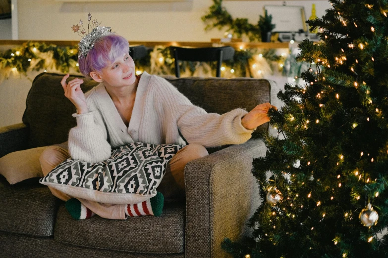 a woman sitting on a couch next to a christmas tree, pexels, lilac hair, queen crown on top of her head, non-binary, wearing pajamas