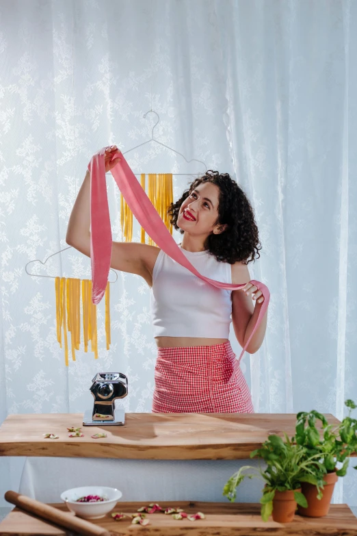 a woman standing in front of a table holding a pink scarf, made of spaghetti, curls on top, hanging, straining