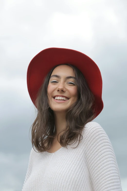 a woman wearing a red hat and a white sweater, fair olive skin, while smiling for a photograph, wearing red, skies