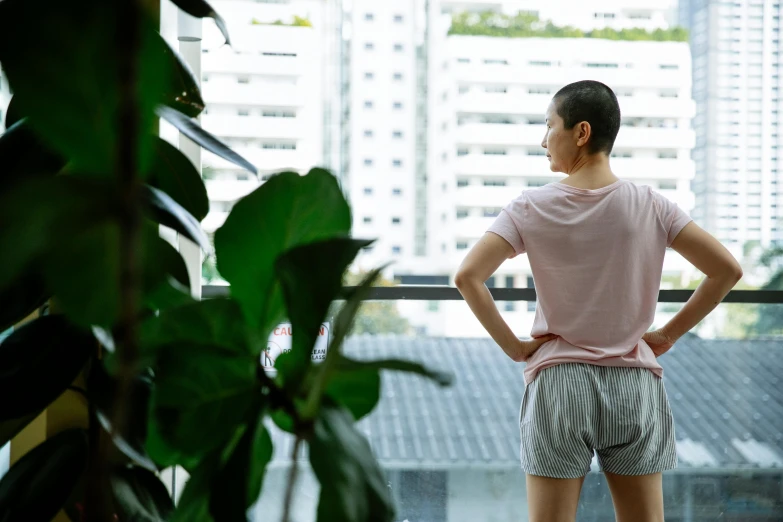 a woman standing in front of a window with her hands on her hips, unsplash, happening, partially bald, plants on balconies, set on singaporean aesthetic, health supporter