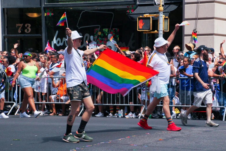 a group of people walking down a street holding a rainbow flag, pexels contest winner, in the macys parade, two men, wearing a tanktop and skirt, holding a white flag
