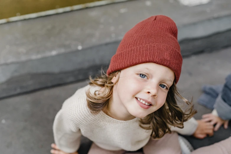 a little girl sitting on top of a skateboard, by Emma Andijewska, pexels contest winner, beanie hat, maroon red, boy with neutral face, smiling down from above