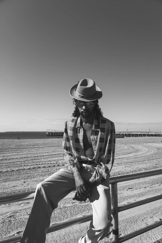 a black and white photo of a man sitting on a fence, by Andrew Stevovich, wearing sunglasses and a hat, on the sand, ashteroth, dressed with long fluent clothes