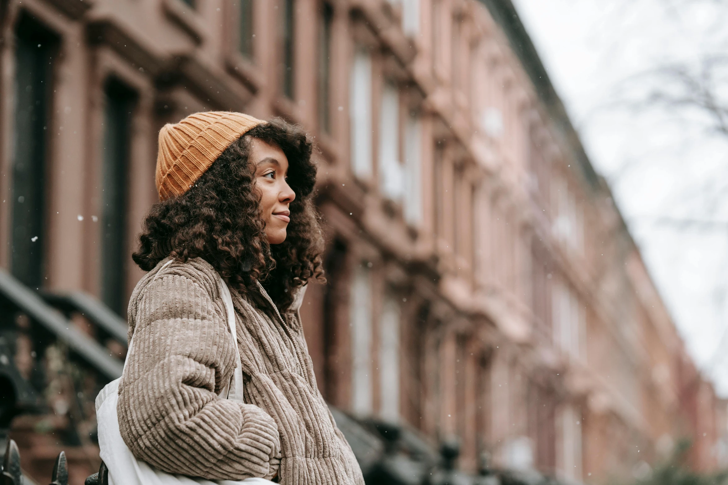 a woman standing on a city street in the snow, trending on pexels, brown curly hair, knitted hat, looking off into the distance, african american girl