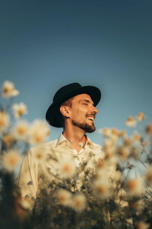 a man with a hat standing in a field of flowers, a picture, by Niko Henrichon, hollywood promotional image, shaven stubble, smiley profile, profile image