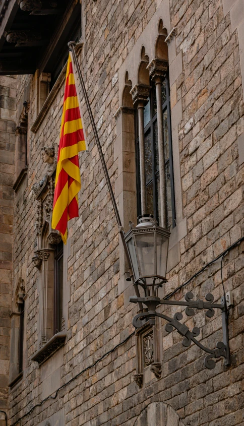 a flag hanging on the side of a brick building, inspired by Modest Urgell, pexels contest winner, renaissance, red and yellow light, square, thumbnail, gothic quarter