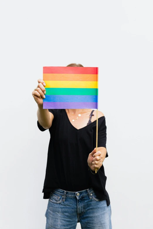 a woman holding a rainbow flag in front of her face, trending on unsplash, holding a wooden staff, with a white background, multiple stories, woman holding sign