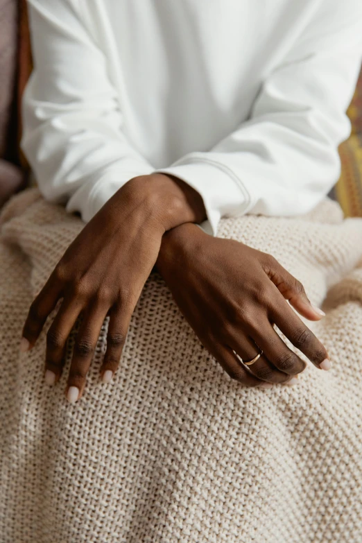 a close up of a person sitting on a couch, by Lily Delissa Joseph, natural hands and arms, gold rings, wearing a white sweater, wearing simple robes