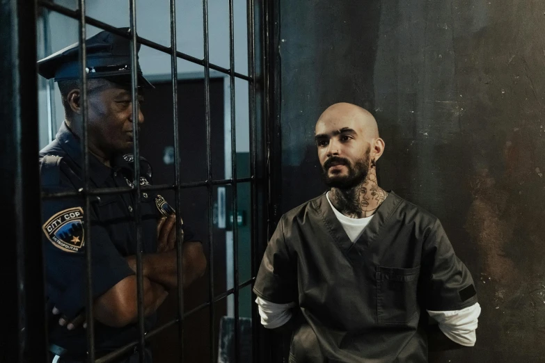 a man sitting in a jail cell next to a police officer, a colorized photo, by Elsa Bleda, pexels contest winner, singer maluma, bald lines, still from a terence malik film, set photo in costume