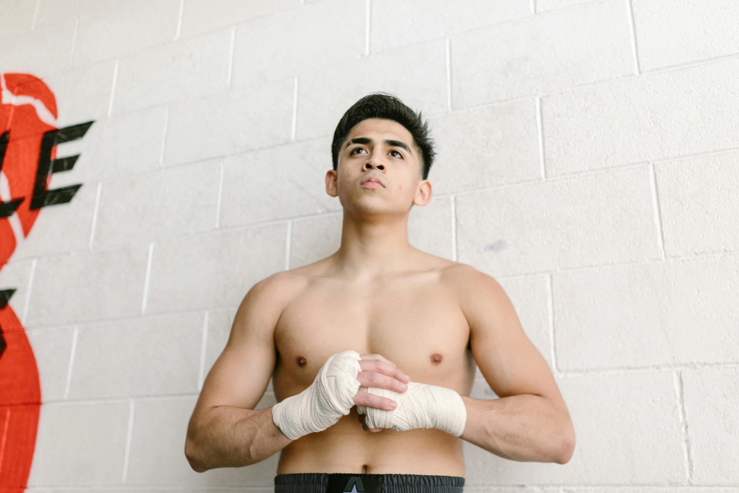 a shirtless young man standing in front of a wall, by Robbie Trevino, pexels contest winner, happening, mma southpaw stance, white bandages on fists, asian male, male teenager