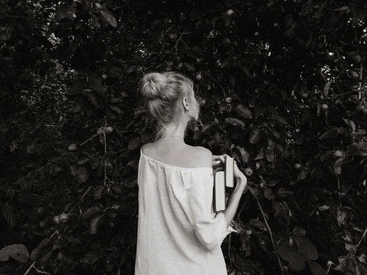 a woman standing in front of a bush holding a book, a black and white photo, inspired by Nell Dorr, pexels contest winner, showing her shoulder from back, young blonde woman, 15081959 21121991 01012000 4k, holding books