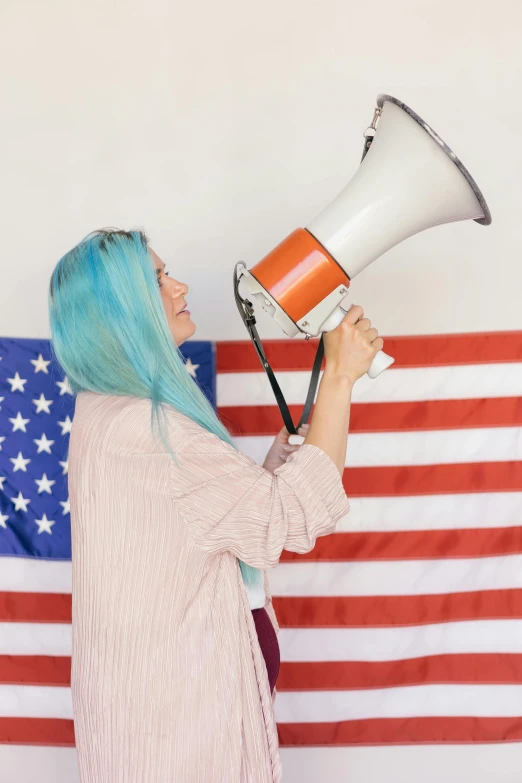 a woman with blue hair holding a megaphone in front of an american flag, a colorized photo, trending on unsplash, long orange hair, subreddit / r / whale, 🚿🗝📝, politics