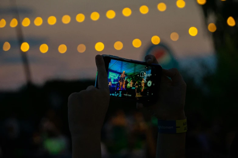 a person holding up a cell phone to take a picture, happening, an outdoor festival stage, firefly lights, looking back at the camera, green concert light