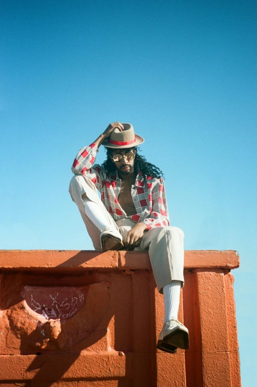 a man sitting on top of a brick wall, an album cover, inspired by Ras Akyem, trending on pexels, white suit and hat, sunfaded, ashteroth, gazing off into the horizon