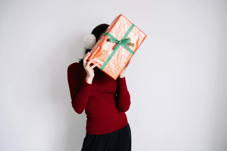 a woman holding a wrapped gift in front of her face, by Alice Mason, pexels contest winner, wearing an orange jumpsuit, with a white background, background image, japanese