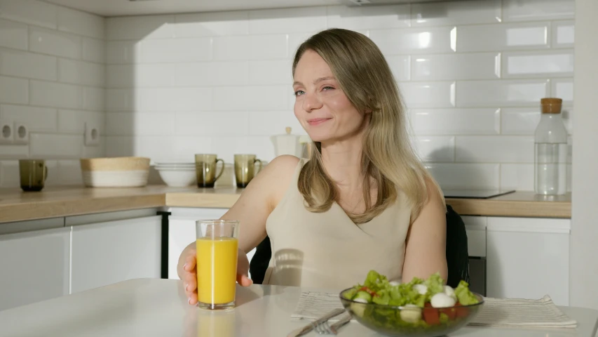 a woman sitting at a table with a glass of orange juice, inspired by Louisa Matthíasdóttir, pexels contest winner, satisfied pose, profile image, on kitchen table, low quality photo