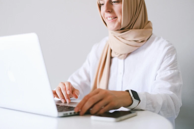 a woman sitting at a table using a laptop computer, inspired by Maryam Hashemi, trending on pexels, hurufiyya, white scarf, plain background, link from zelda using computer, professional closeup photo