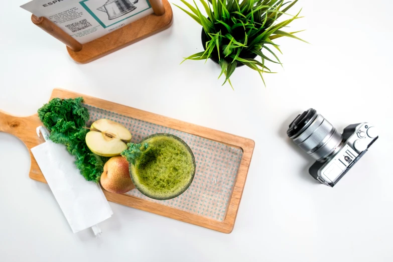 a wooden cutting board topped with an apple and green smoothie, a picture, inspired by Art Green, unsplash, hasselblad photo, green square, official product image, on a white table