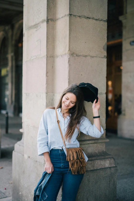 a woman leaning against a pillar with a hat on her head, by Julia Pishtar, trending on unsplash, carrying a saddle bag, blue shirt, brunette, hair fanned around