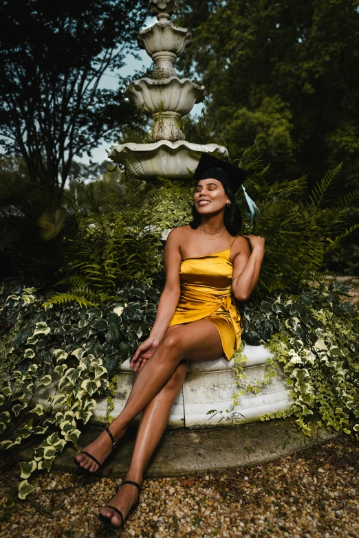 a woman in a yellow dress sitting in front of a fountain, a photo, by Carey Morris, unsplash, graduation photo, with brown skin, lush surroundings, awards winning