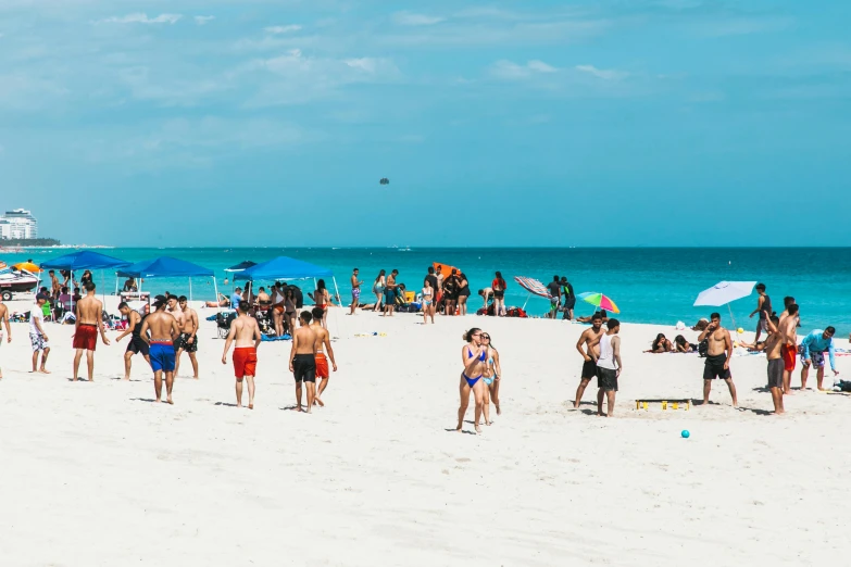 a group of people standing on top of a sandy beach, by Robbie Trevino, miami beach, people with umbrellas, dua lipa, on a sunny beach