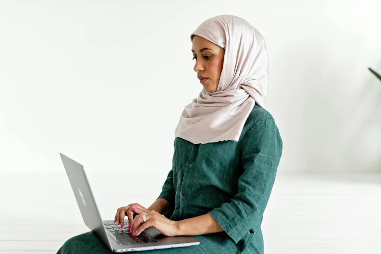 a woman sitting on the floor using a laptop, inspired by Maryam Hashemi, hurufiyya, wearing green clothing, wearing a scarf, set against a white background, on a desk