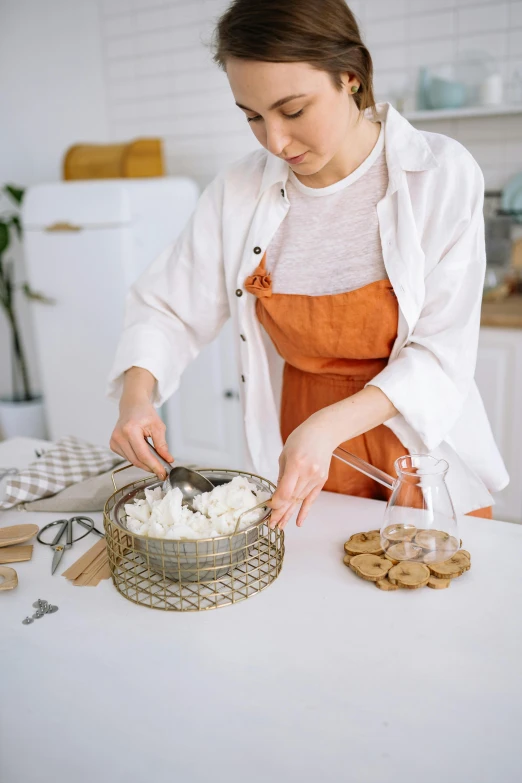 a woman in an orange apron is making a cake, inspired by Yukimasa Ida, pexels contest winner, cream and white color scheme, curated collections, metal kitchen utensils, white