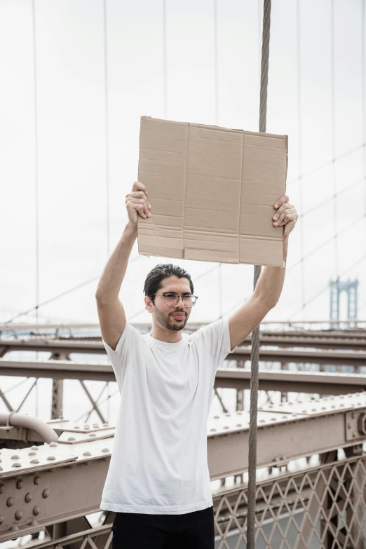 a man holding up a sign on top of a bridge, inspired by Christo, pexels contest winner, serial art, adam driver, cut out of cardboard, brooklyn, delivering parsel box