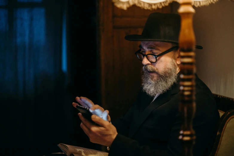 a man sitting in a chair using a cell phone, spell casting wizard, wearing a fedora, orthodox, wearing black rimmed glasses