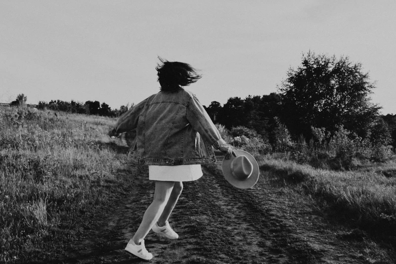 a woman walking down a dirt road holding a hat, a black and white photo, pexels, girl running, carefree, wearing a jeans jackets, girl in white dress dancing