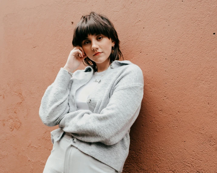 a woman leaning against a wall talking on a cell phone, an album cover, inspired by Ion Andreescu, trending on pexels, grey sweater, confident pose, bangs, white clothing