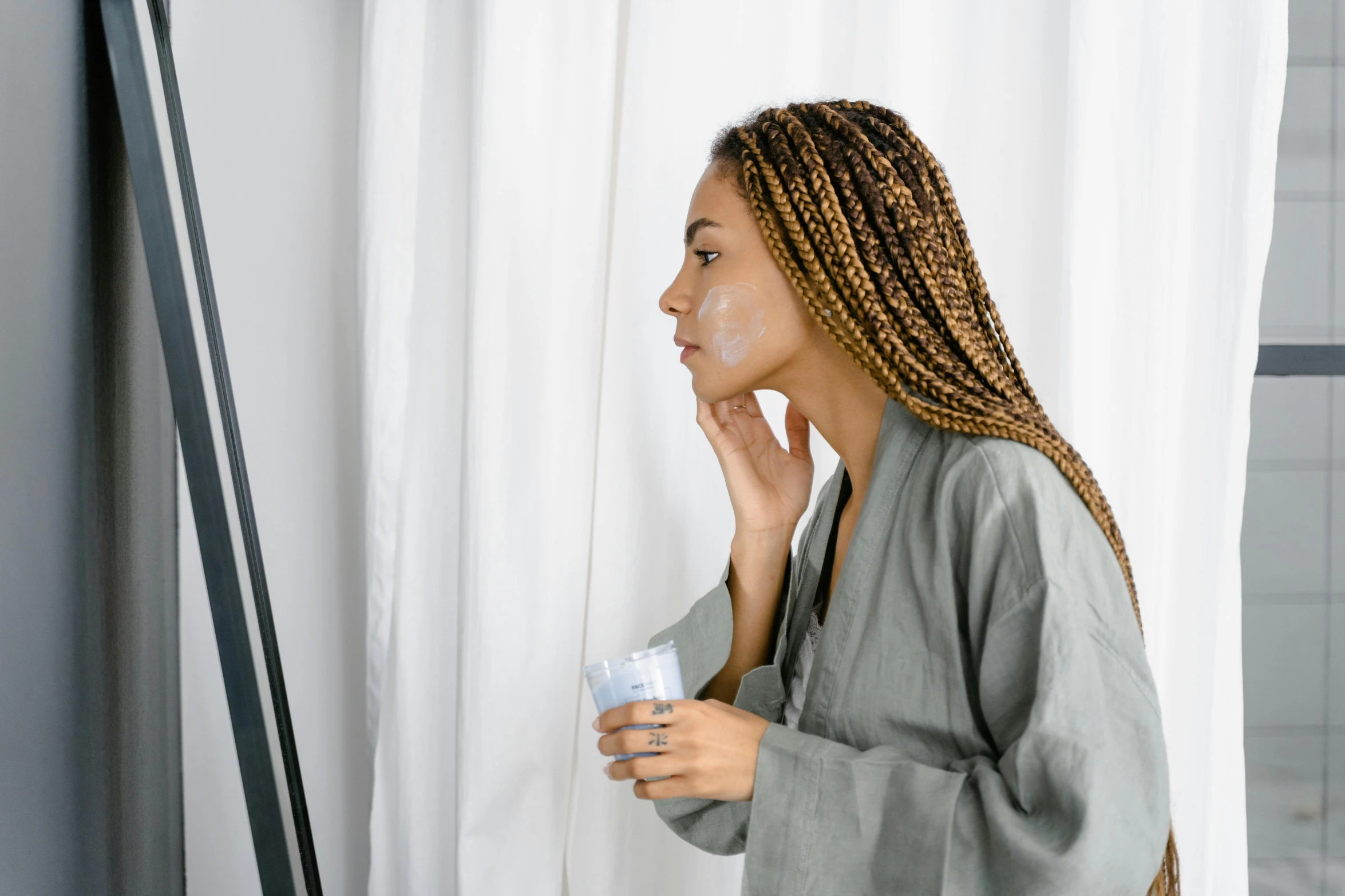 a woman standing in front of a window holding a toothbrush, a hyperrealistic painting, by Emma Andijewska, trending on pexels, black scars on her face, wearing a grey robe, skincare, looking in mirror