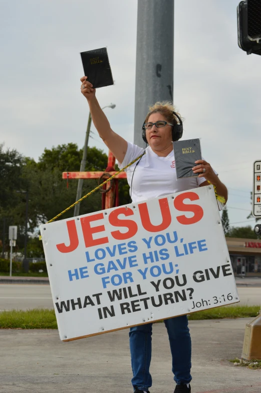 a woman holding a sign that says jesus loves you, he gave his life for you what will you give in return?, trending on reddit, florida man, ap news photo, trans rights, roadside