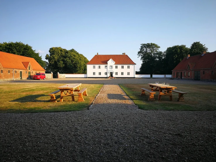 a couple of picnic tables sitting on top of a gravel field, a picture, by Jesper Knudsen, heidelberg school, mansion, oland, white buildings with red roofs, a middle-shot from front