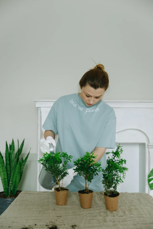 a woman is watering plants on a table, wearing a light blue shirt, with a tall tree, plain background