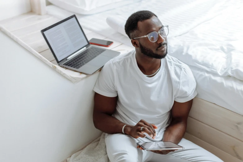 a man sitting on a bed in front of a laptop, trending on pexels, with brown skin, tech glasses, sleek white, an all white human