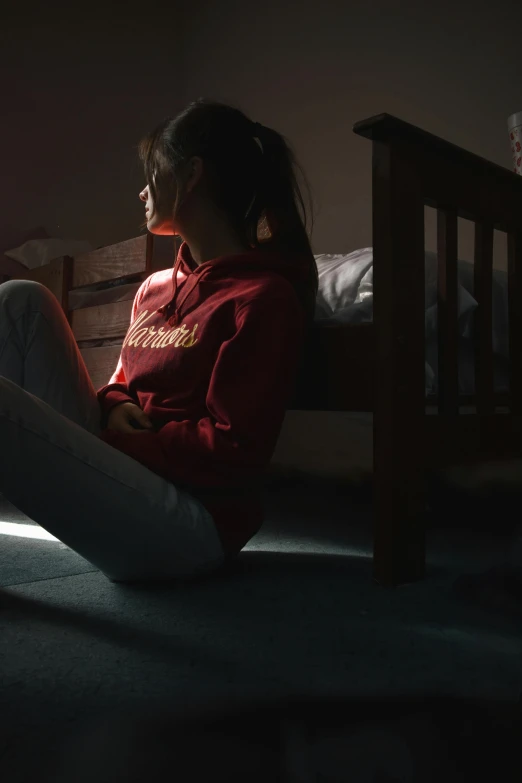 a woman sitting on a bed in a dark room, wearing a red hoodie, teenager hangout spot, late afternoon lighting, promo image