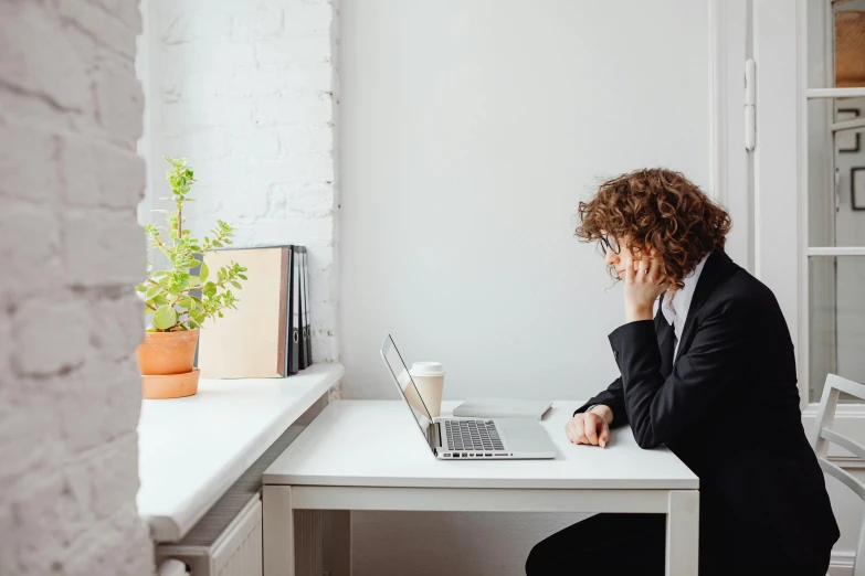 a person sitting at a table with a laptop, by Nicolette Macnamara, trending on pexels, figuration libre, tired half closed, in a white room, woman in black business suit, next to a plant