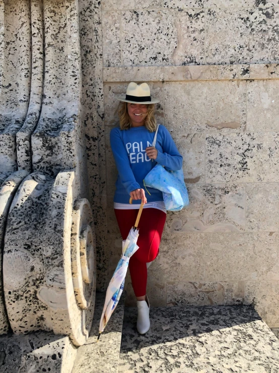 a woman leaning against a wall with an umbrella, a photo, by Pamela Drew, pexels contest winner, red and blue garments, wears a destroyed hat, near a stone gate, fun pose