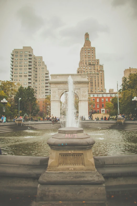 a fountain in the middle of a city with tall buildings in the background, archway, new york backdrop, square, college