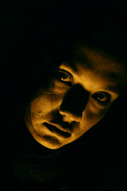 a man staring at the camera in the dark, glowing yellow face, with haunted eyes and dark hair, ( ( theatrical ) ), young woman's face