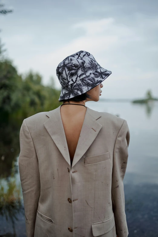 a woman standing in front of a body of water, bucket hat, wearing fashion suit, trees in the background, with his back turned