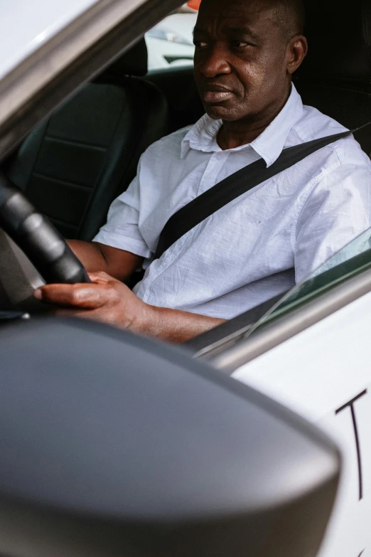 a man sitting in the drivers seat of a car, wearing white shirt, dark skinned, concentration, over the shoulder