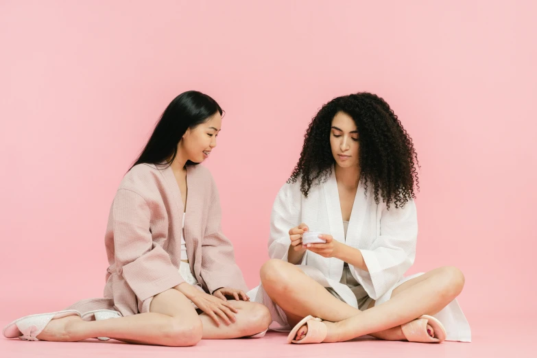 two women sitting next to each other on a pink surface, trending on pexels, wearing a white robe, avatar image, manicured, manuka