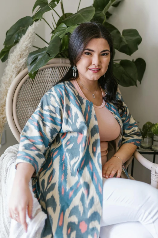 a woman sitting in a chair next to a potted plant, a portrait, by Basuki Abdullah, hurufiyya, loose - fitting blouses, cardigan, white and teal garment, trending photo