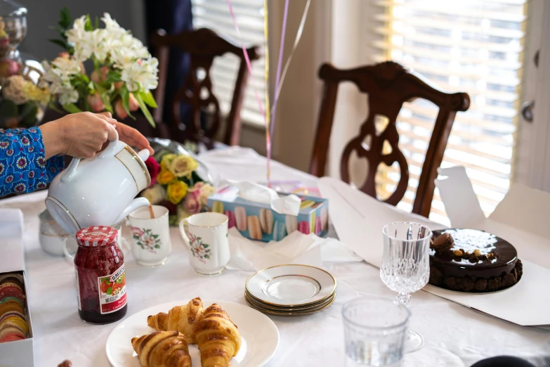 a woman pours tea over a plate of croissants, a still life, by Liza Donnelly, pexels contest winner, at a birthday party, french provincial furniture, dslr photo of a vase on a table, party balloons