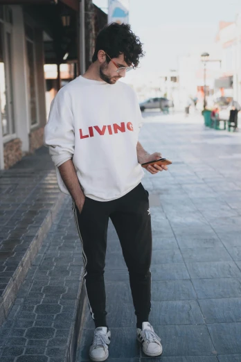 a man standing on a sidewalk looking at his cell phone, an album cover, trending on pexels, white sweater, living, outlive streetwear collection, full - body - front - shot