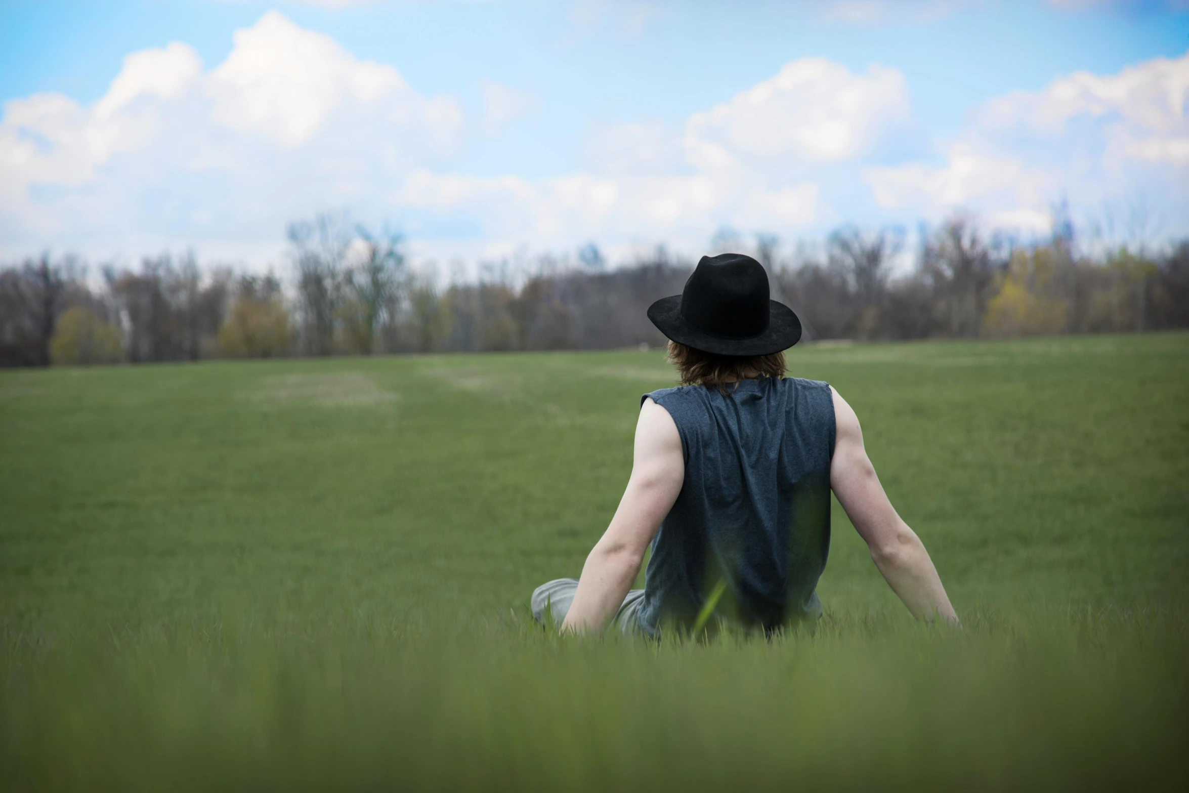 a person sitting in a field with a hat on, pexels, wearing a black leather vest, green field, his back is turned, heath clifford