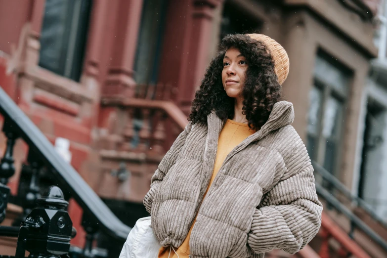 a woman standing in front of a brick building, trending on pexels, happening, puffer jacket, harlem, brown curly hair, wearing an oversized sweater