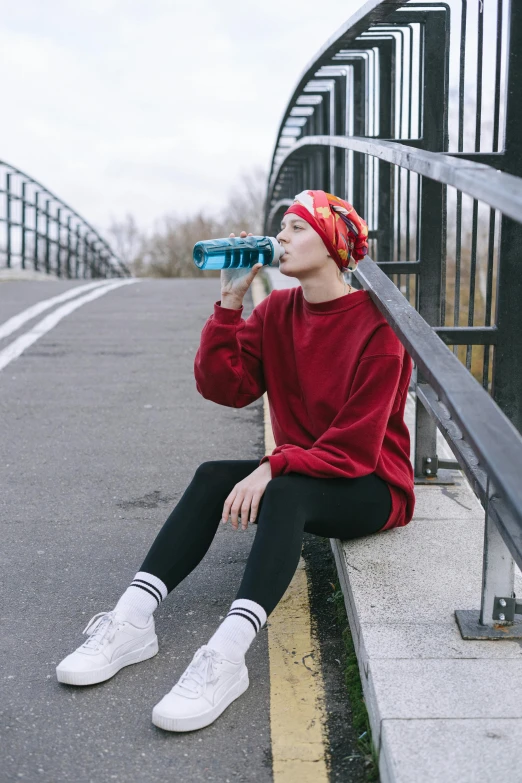 a woman sitting on the side of a road drinking water, pexels contest winner, wearing teal beanie, non binary model, energy drink, wearing a red turtleneck sweater